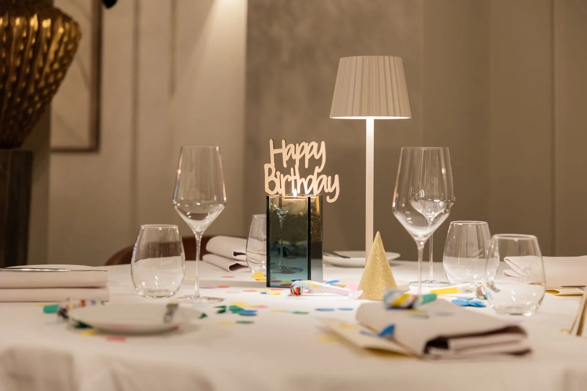 Gedekte tafel met opschrift Happy Birthday, confetti, feesthoedje, toeters en tafellamp.