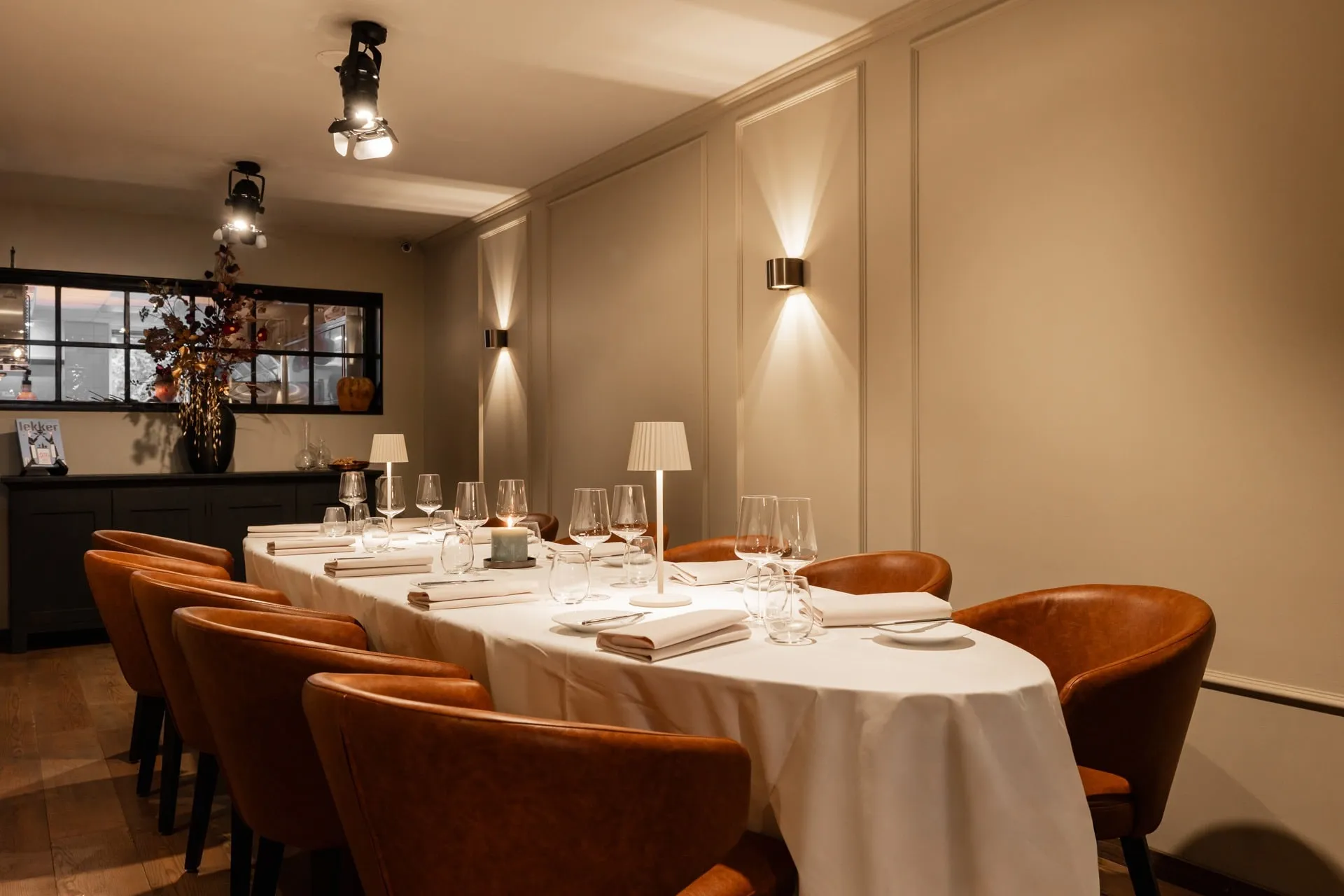 Lange gedekte tafel met wit tafelkleed, leren stoelen, wijnglazen en tafellampjes